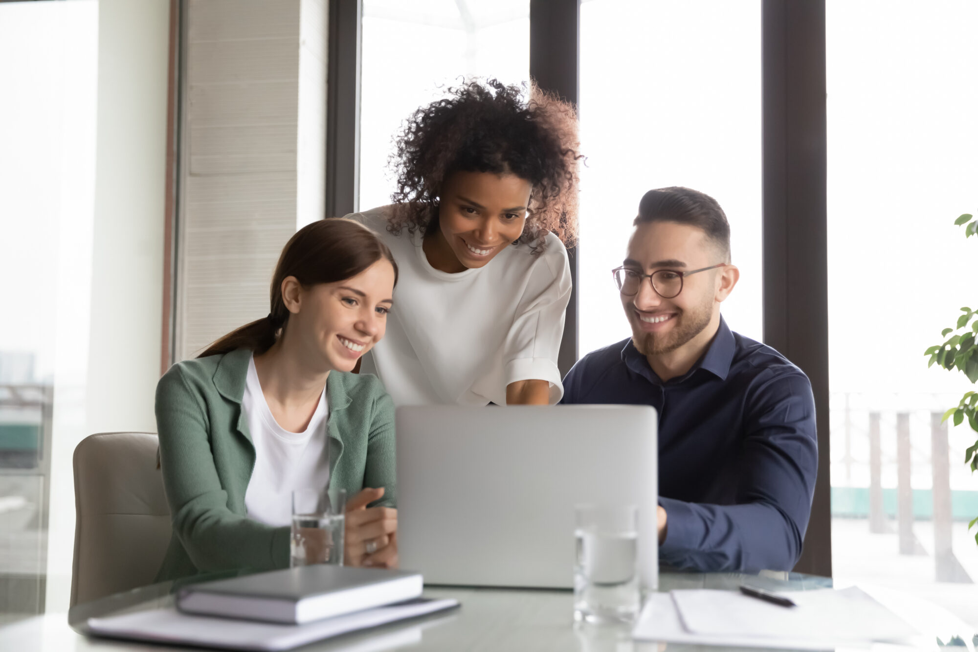 Three happy diverse colleagues working on project together, using laptop, sitting at table in office, discussing strategy, brainstorming, smiling African American businesswoman training staff